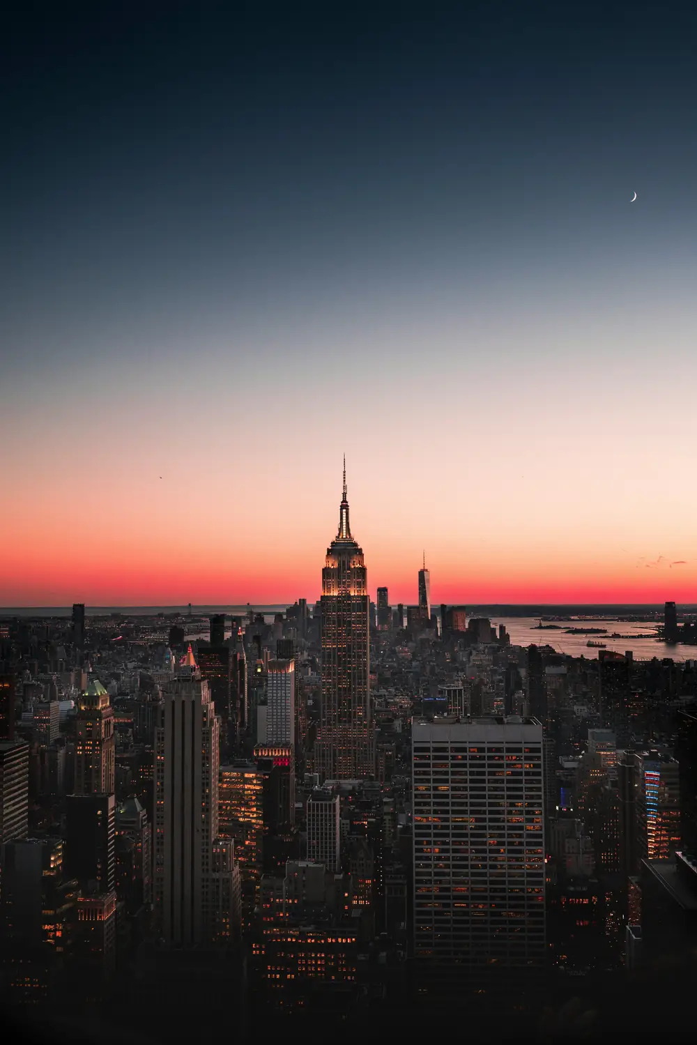 Midtown Manhattan at dusk, the Empire State Building lit against a red horizon and a crescent moon.