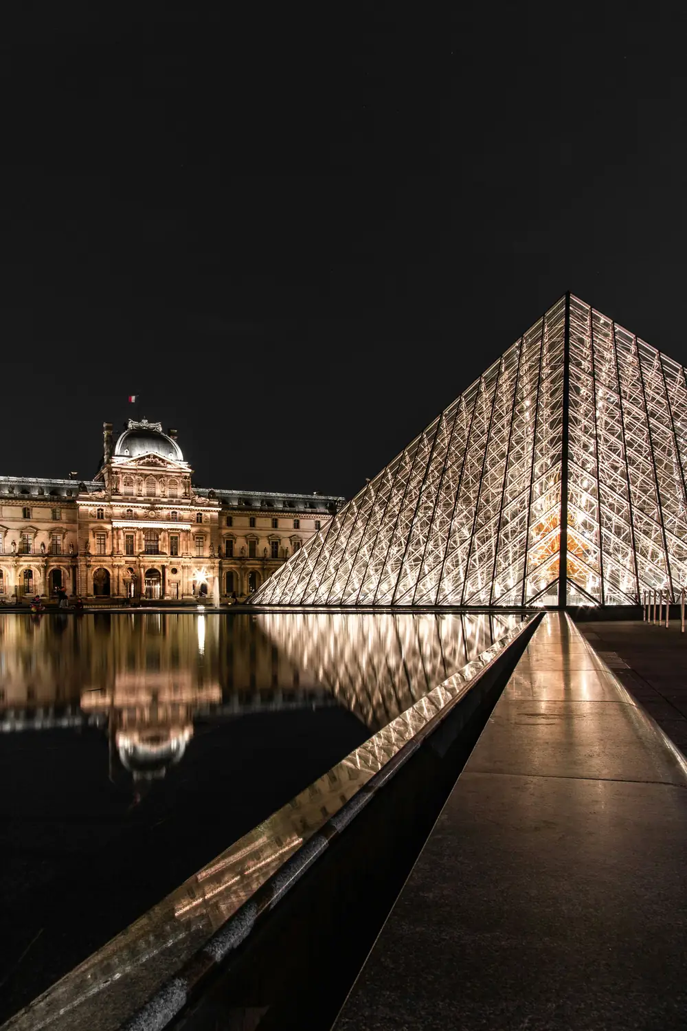 The Louvre pyramid lit at night, reflected in the still water of the courtyard pool.